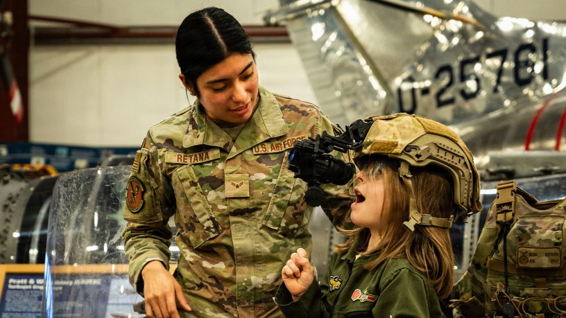 U.S. Air National Guard Airman 1st Class Glini Retana, a security forces specialist with the 103d Security Forces Squadron, helps a child try on a specialized headset at a Women Take Flight event on March 14, 2025, Windsor Locks, Connecticut. Multiple volunteers from the 103d supported this event with hands-on activities and speaking opportunities.