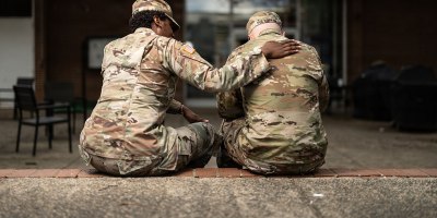 A U.S. Army soldier places a comforting hand on the shoulder of a fellow service member as the two sit together outside a building on what appears to be a military installation. The gesture reflects the importance of peer support and mental health awareness within the armed forces.