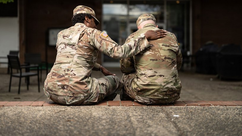 A U.S. Army soldier places a comforting hand on the shoulder of a fellow service member as the two sit together outside a building on what appears to be a military installation. The gesture reflects the importance of peer support and mental health awareness within the armed forces.